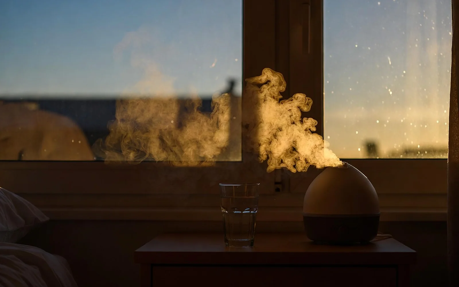 A dim, warm bedroom in the evening, a bedside lamp glowing amber over an open paper book and a mug, the window deep blue with dusk.