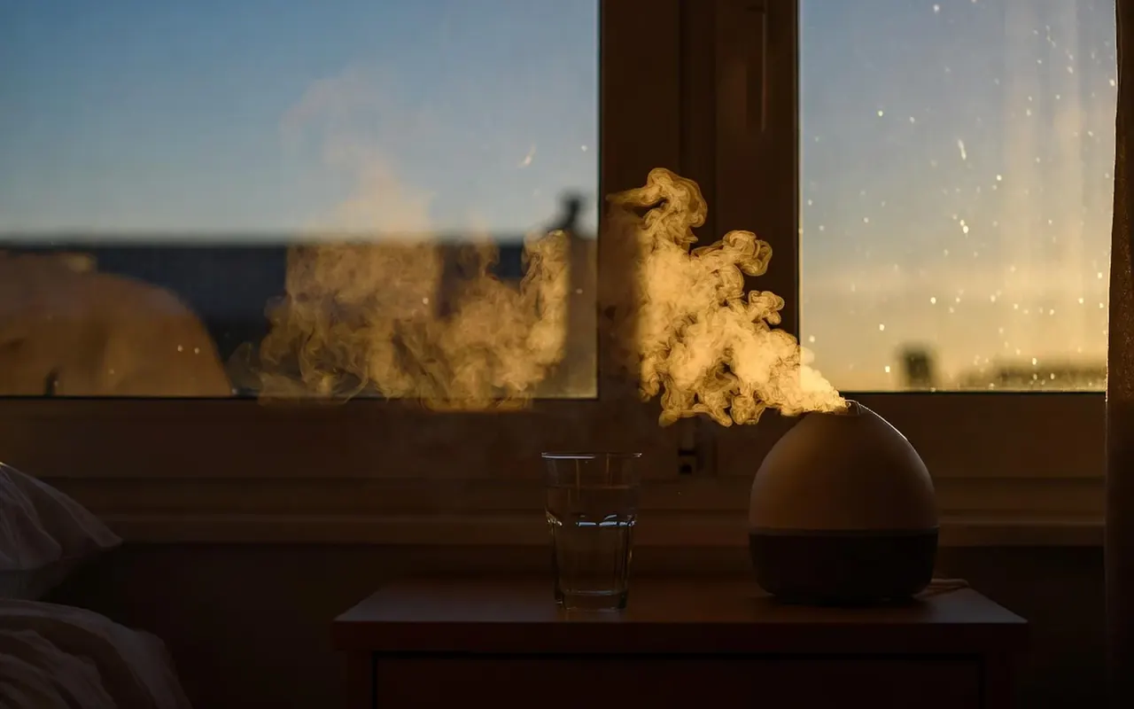 A humidifier on a bedside table sending fine mist into warm evening light, beside a glass of water at the window.