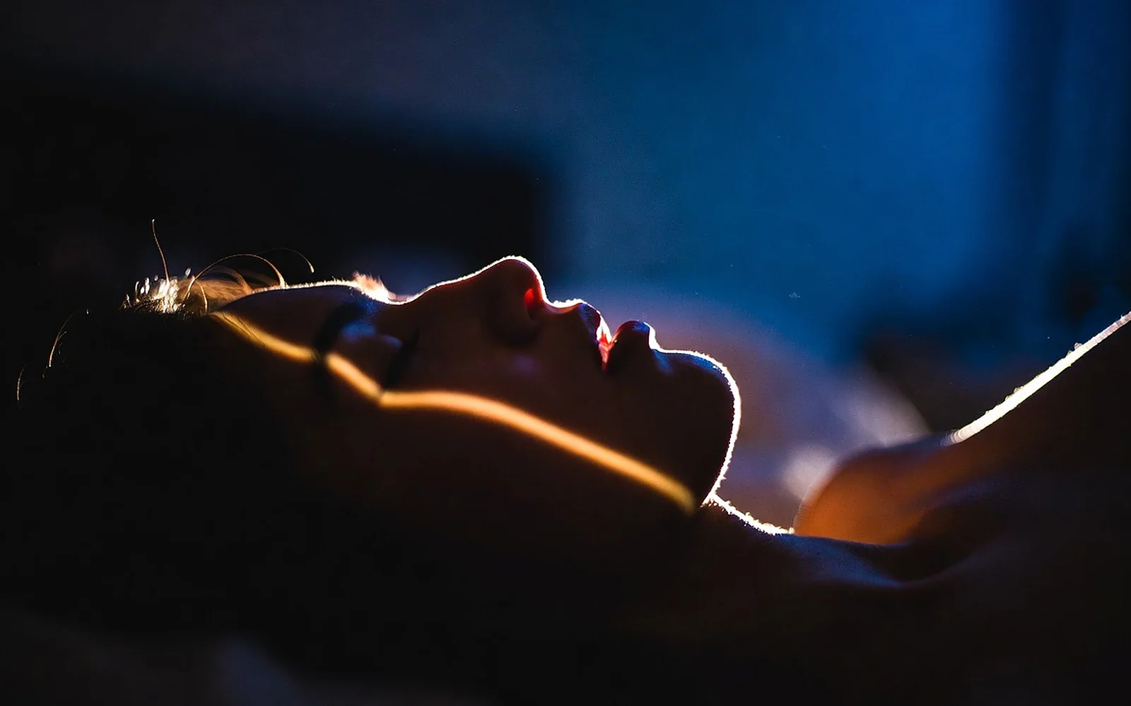 A dark bedroom at night, a person asleep on their side under warm linens, a sliver of moonlight tracing slow, even shadows.