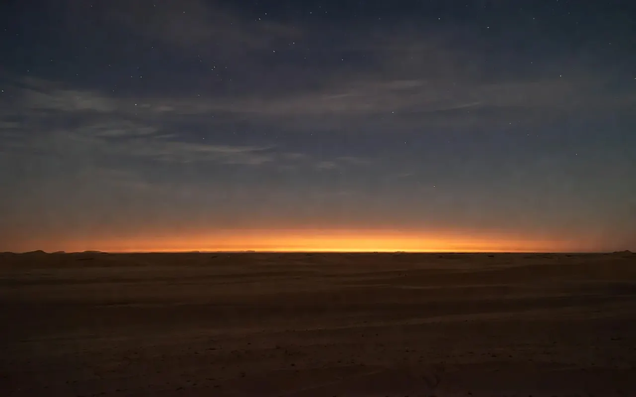 A single warm lantern glowing on dark desert sand at dusk, a thin band of ember light along the horizon beneath early stars.