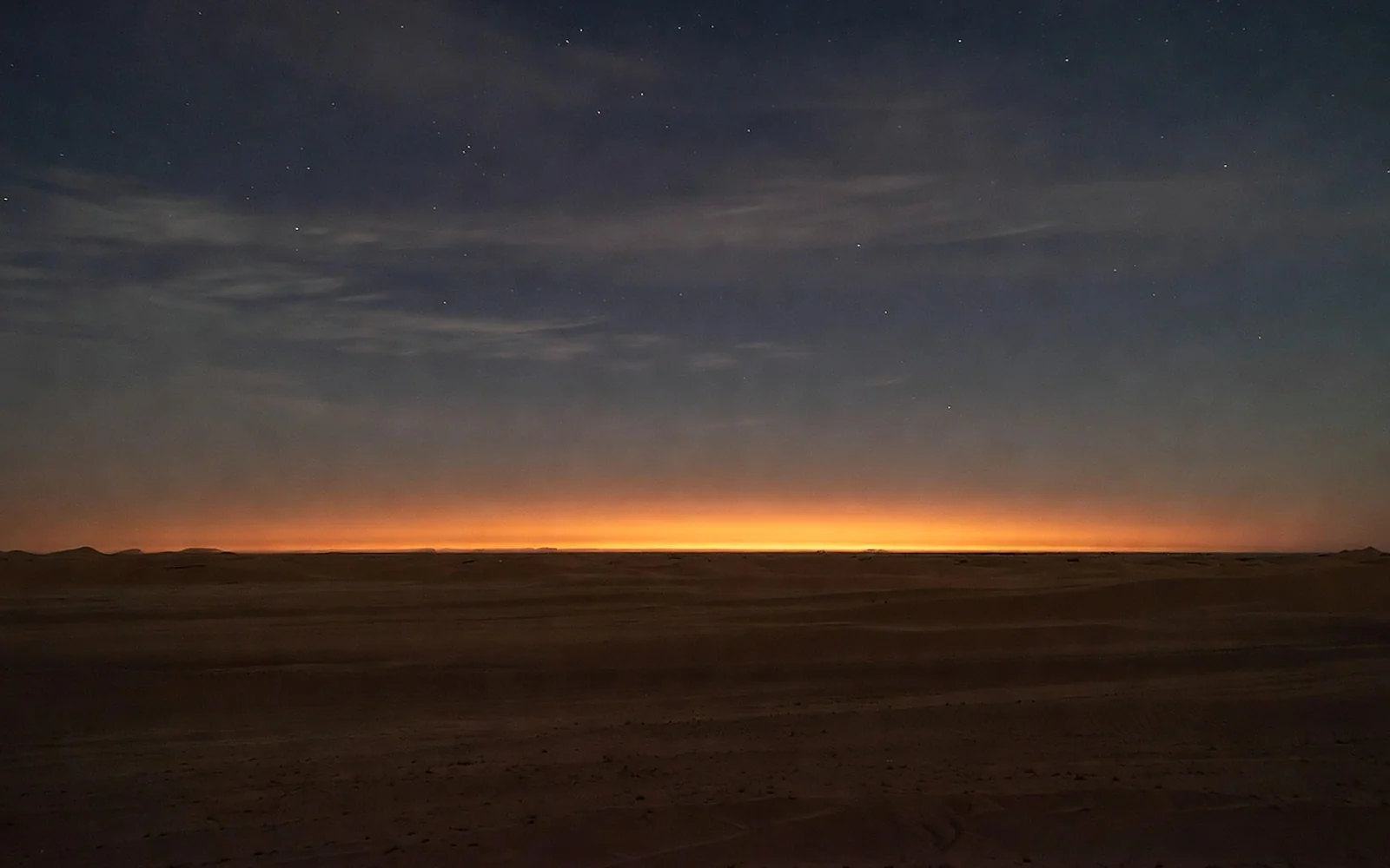 A single warm lantern glowing on dark desert sand at dusk, a thin band of ember light along the horizon beneath early stars.