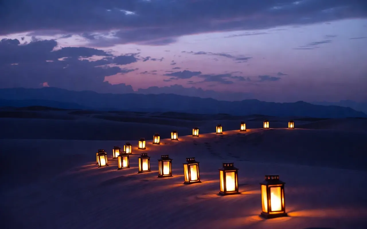 A line of small glowing lanterns crossing desert sand at dusk, rising toward a distant mountain horizon.